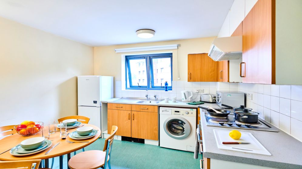 A kitchen with a round table that has been set for four. There is an electric hob, washing machine, fridge freezer, microwave, toaster and kettle.